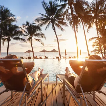 A picture of two persons sitting relaxed enjoying a sunset in front of a pool, close to a beach